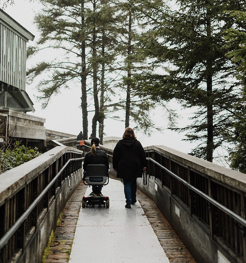 Person in a motorized wheelchair with another person walking along a boardwalk on a cloudy day