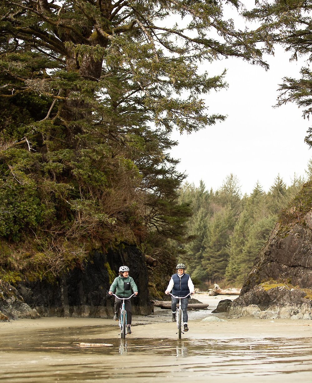 Two people cycling on Long Beach  with tidal rocks and trees in the background