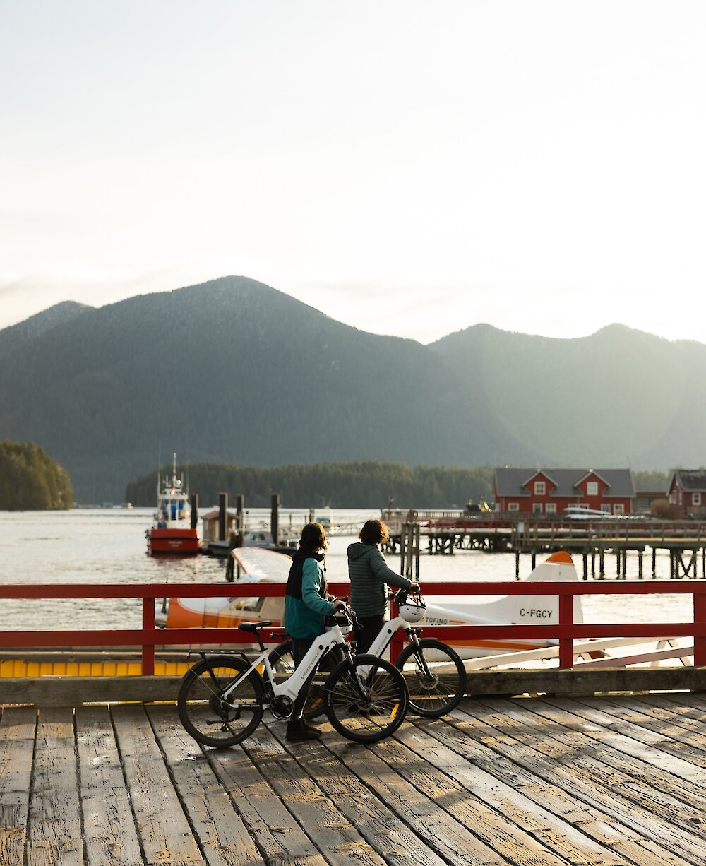 Two people walking their bicycles on a dock with harbour and mountain views