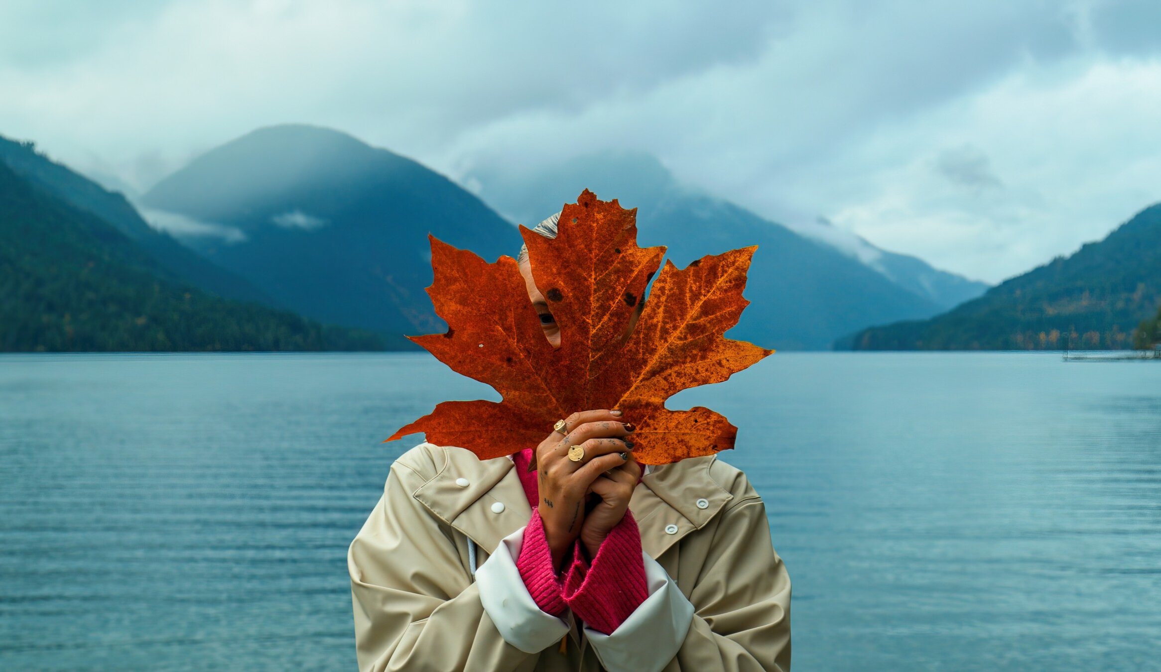 Woman holding giant red maple leaf in front of her face with backfrop of lake.