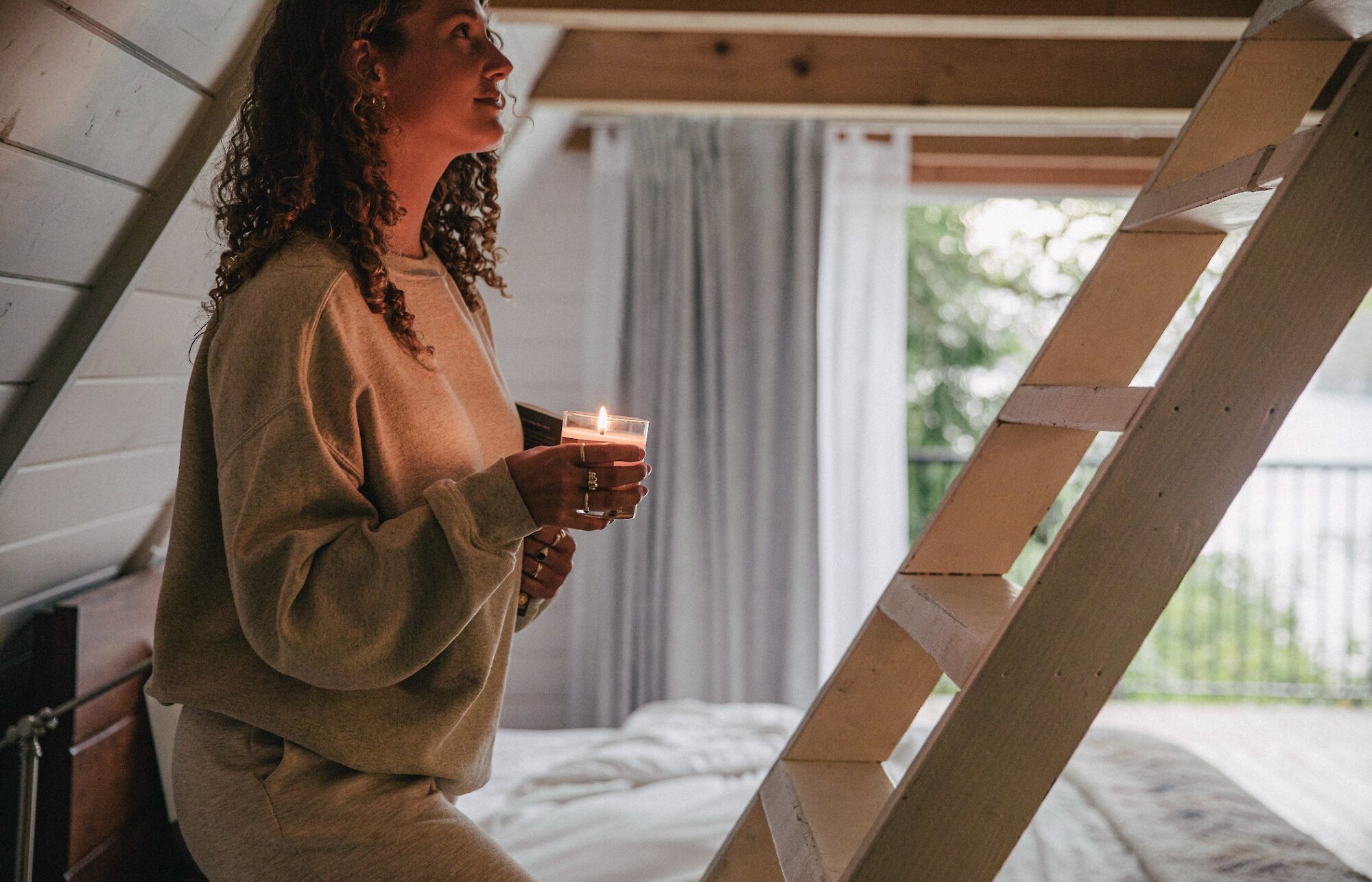 Woman with curly hair holding a candle and climbing up a slanted wooden ladder with ocean views in background