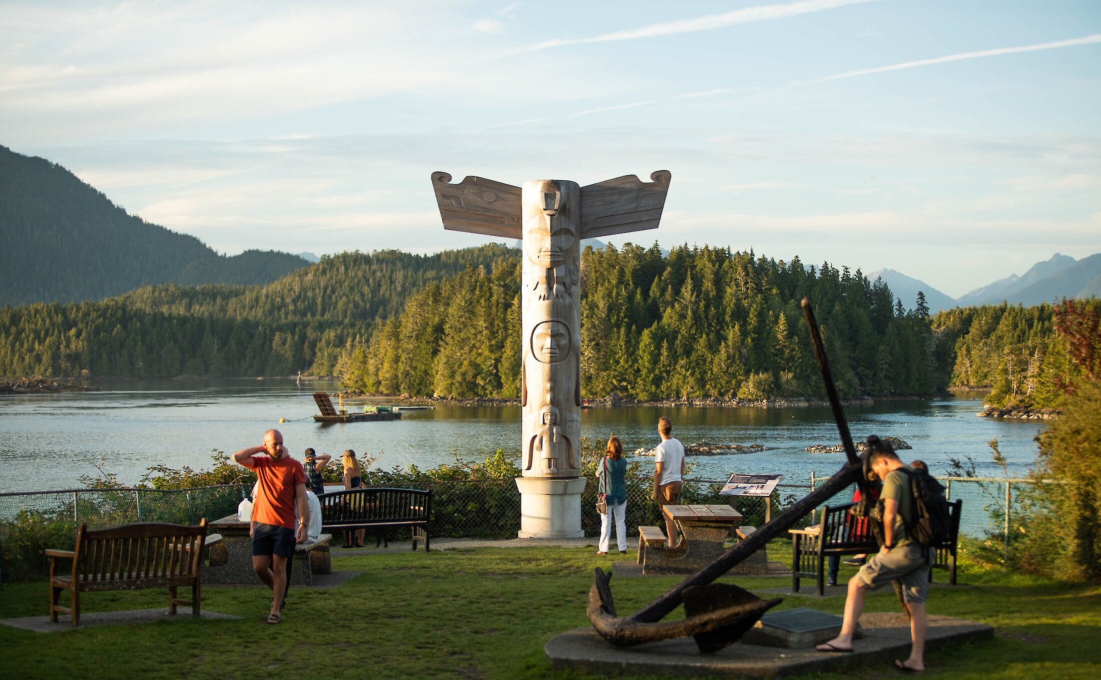 Person looking at a large anchor in a park with a totem pole and view of the water and working Tofino Harbour in the back