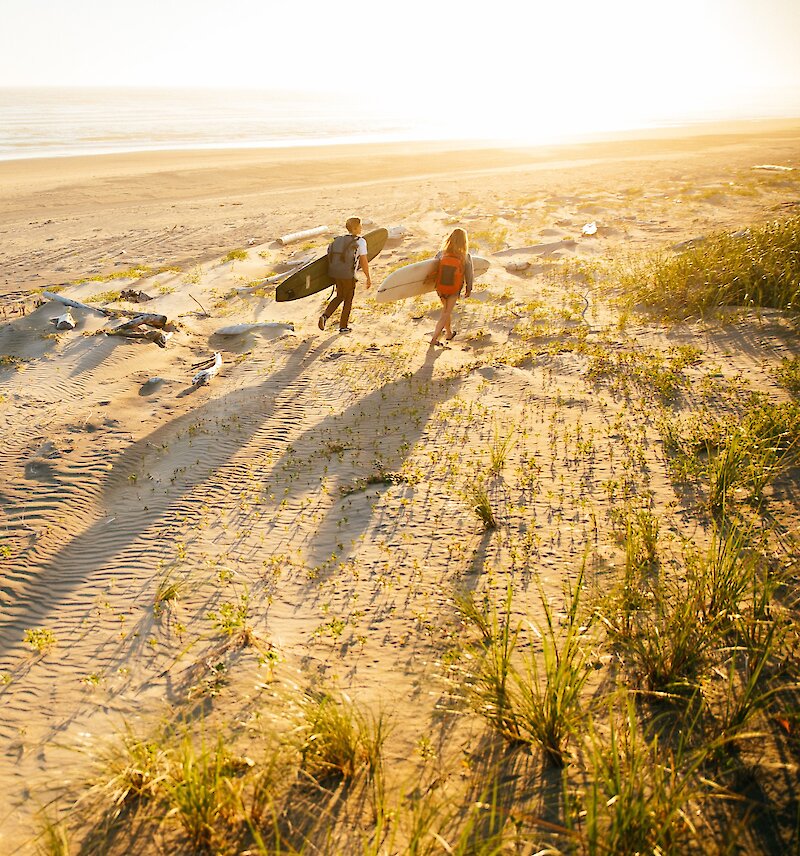 Two people walking along the dunes at Wickaninnish Beach with surfboards in hand