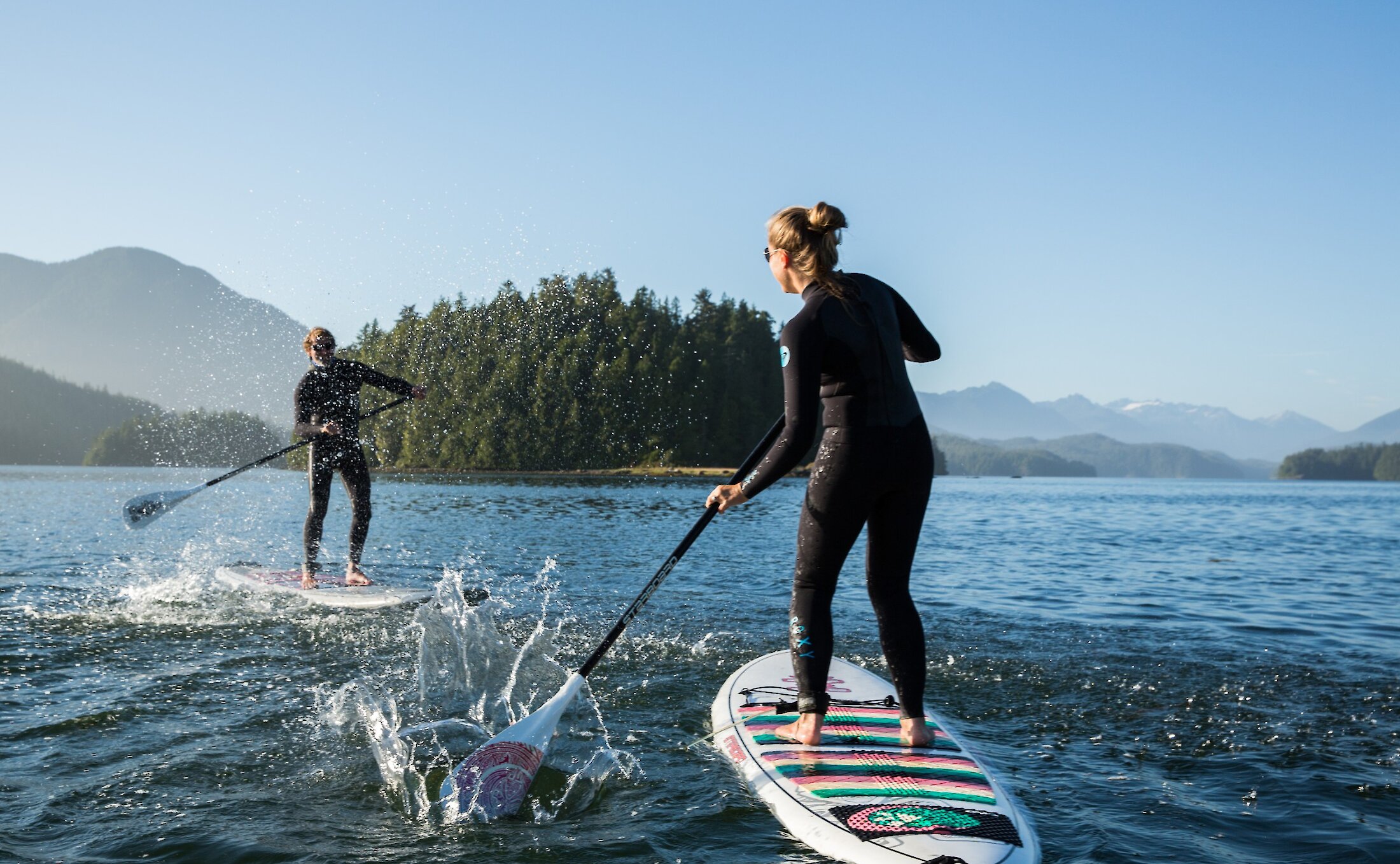 woman and man in wetsuits on paddleboards and splashing water at each other