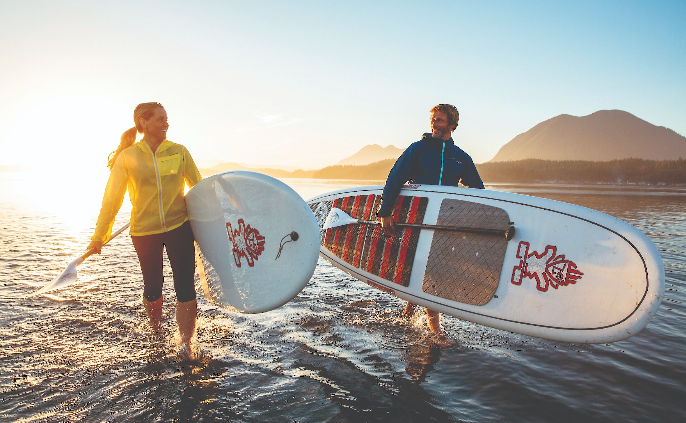 woman and man in jackets holding paddlebpards and walking out of the ocean at sunset