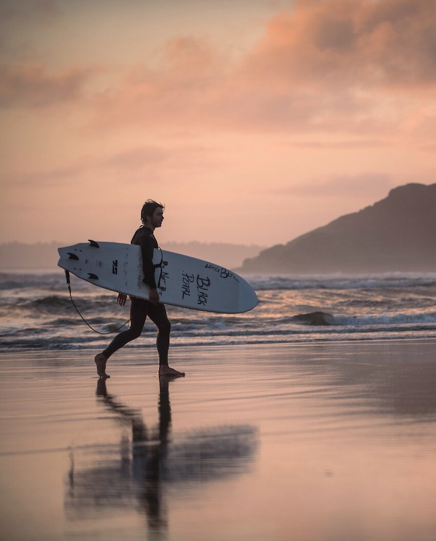 Surfer walking on beach holding surfboard at pink sunset