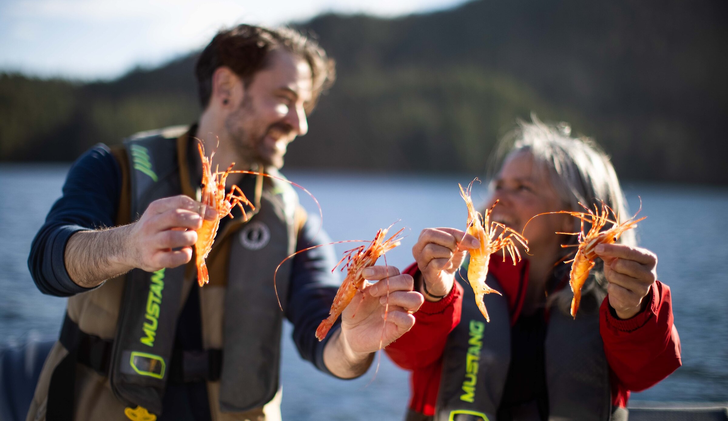 Man in his 30s with beard and woman with grey hair in life jackets, smiling at each other and holding spot prawns