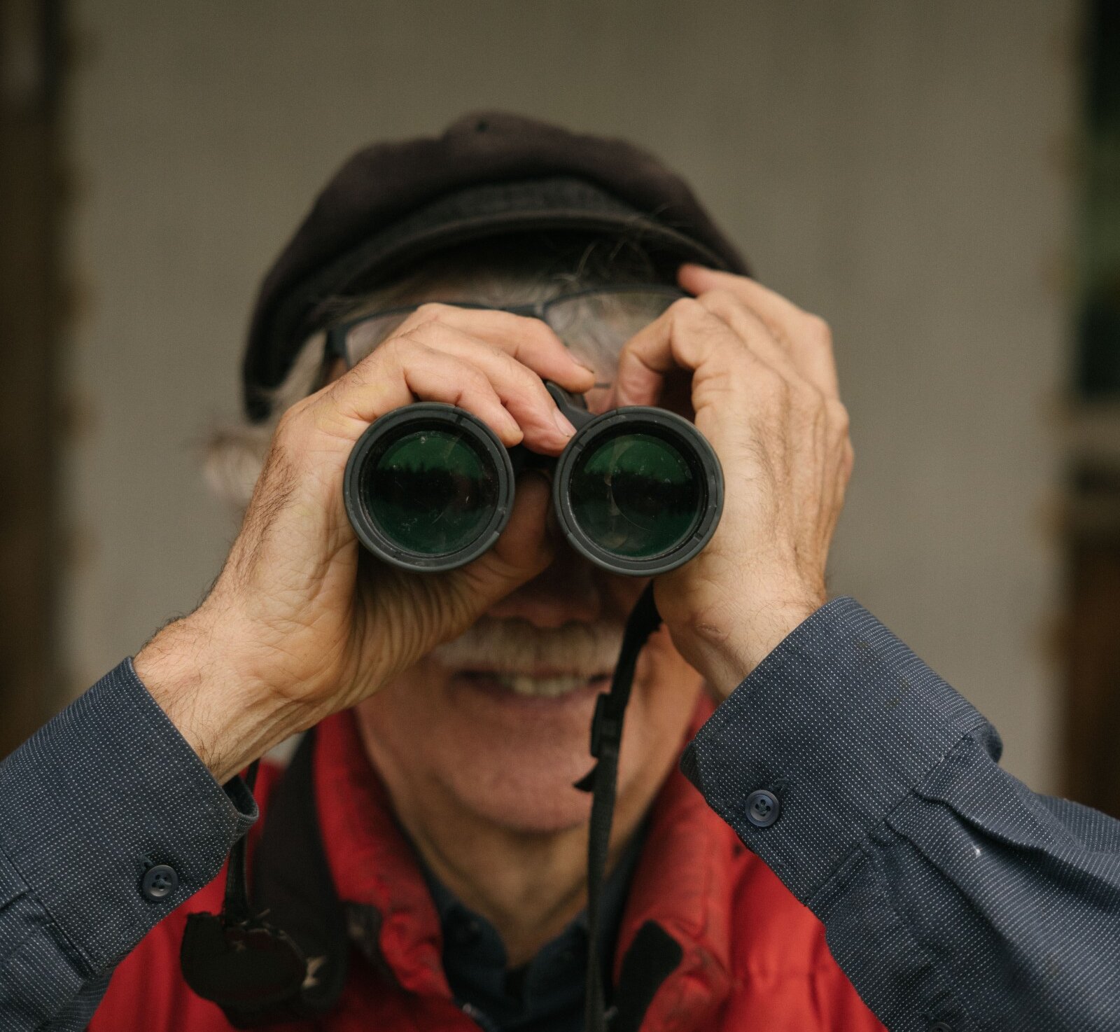 Man in red vest and hat facing camera through binoculars