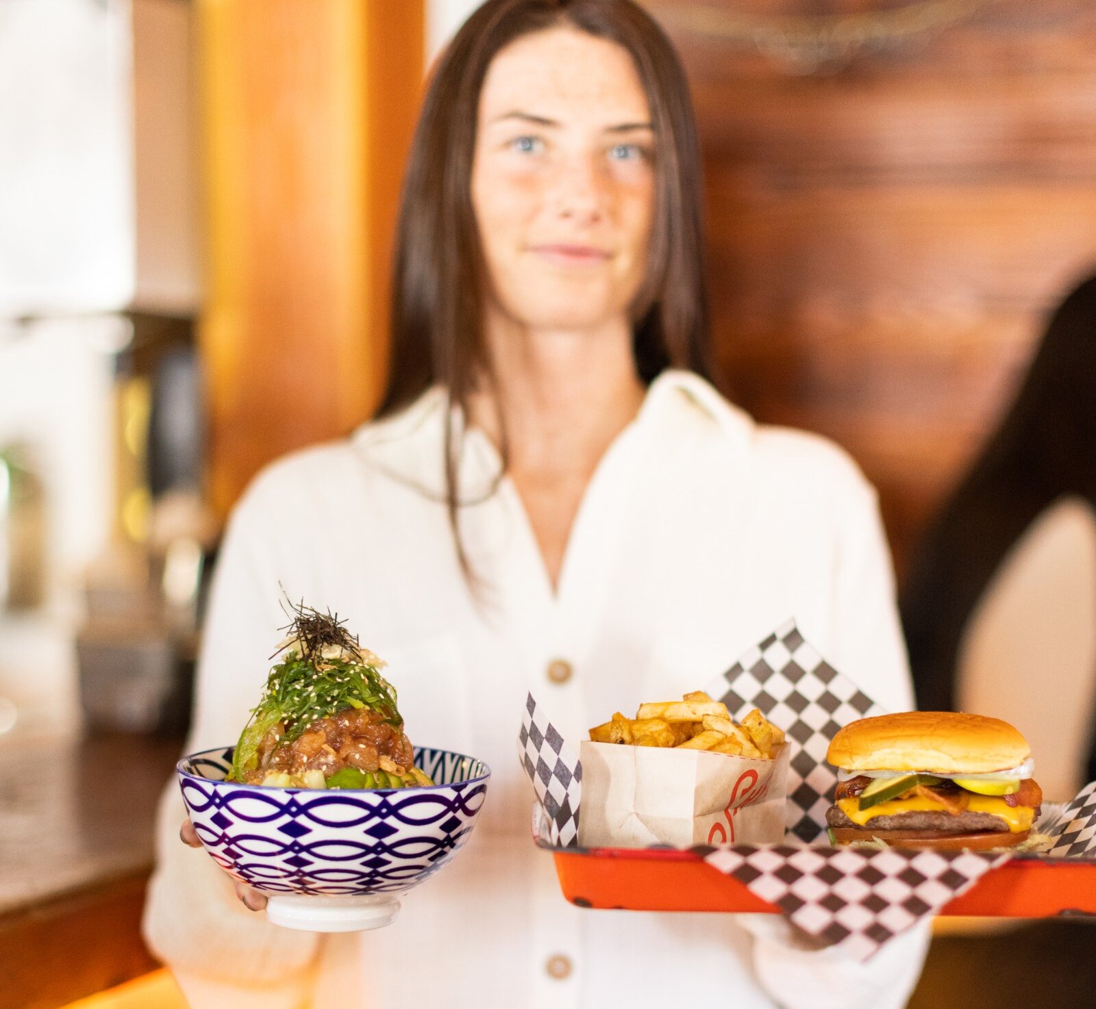 Woman with long hair and freckles holding a poke bowl and a tray with a burger and fries.