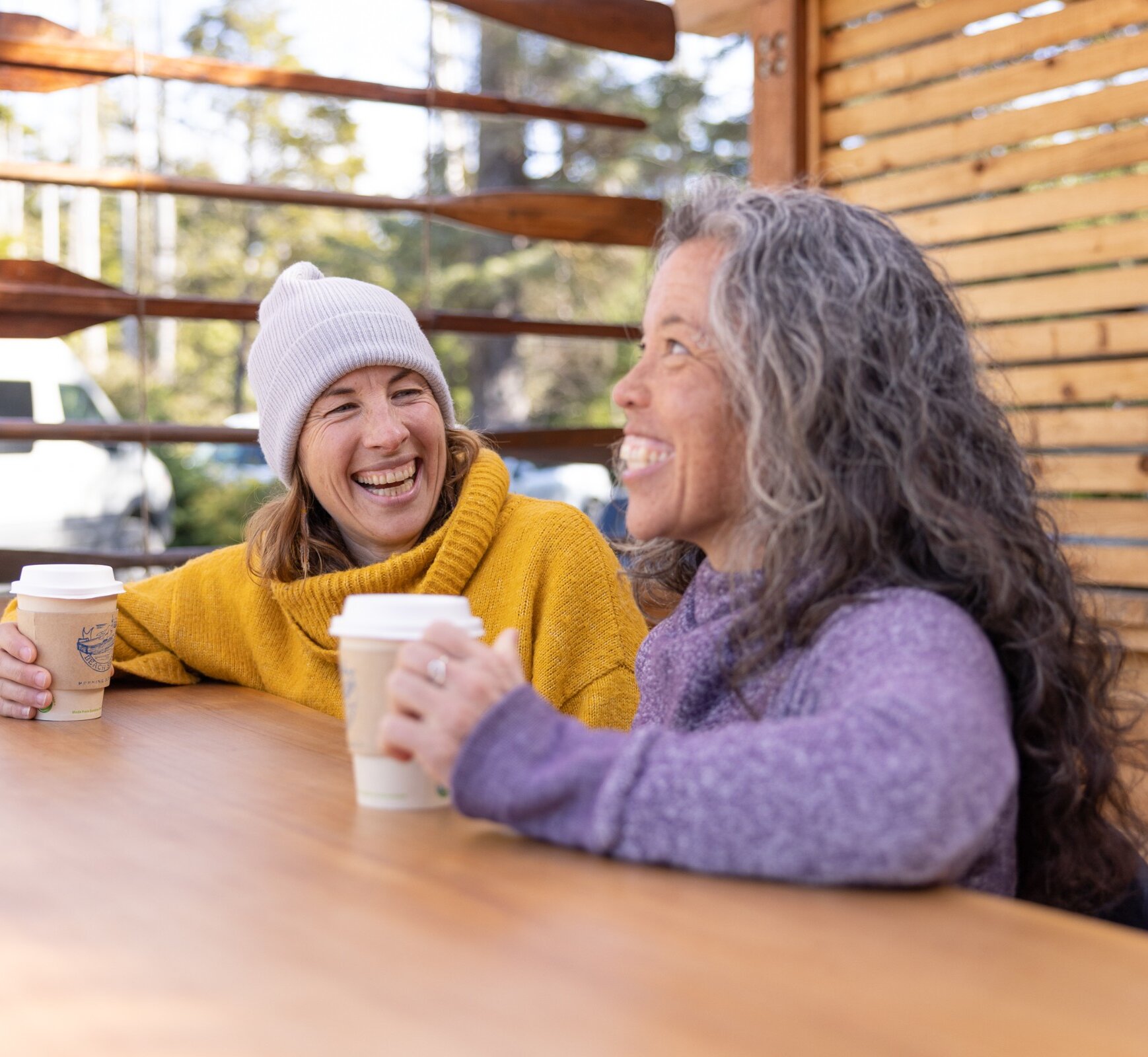 One caucasian woman and a mixed-race woman laughing and drinking at an outside cafe.