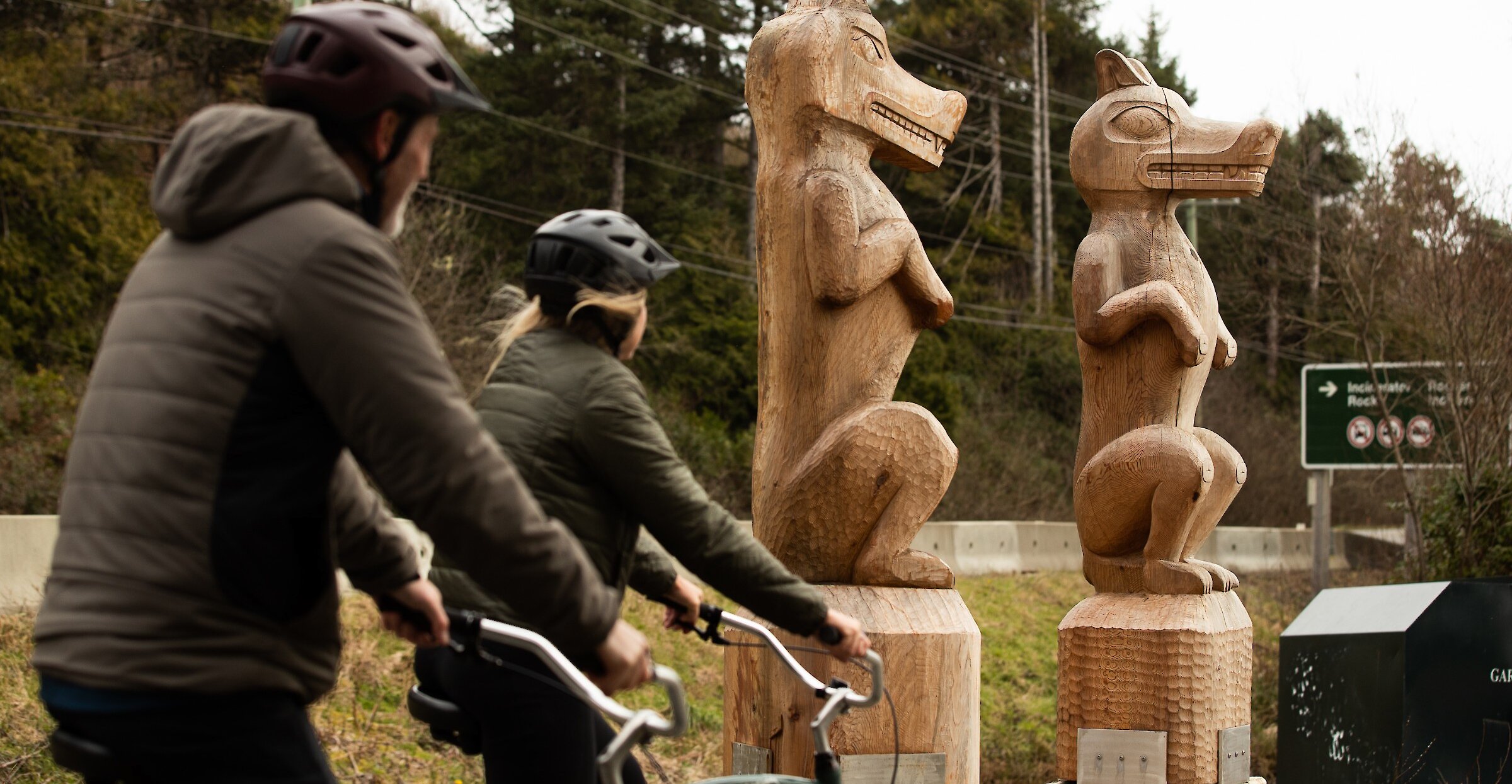 Two people on bikes looking at two wolf welcome figure carvings at Long Beach in Pacific Rim National Park Reserve
