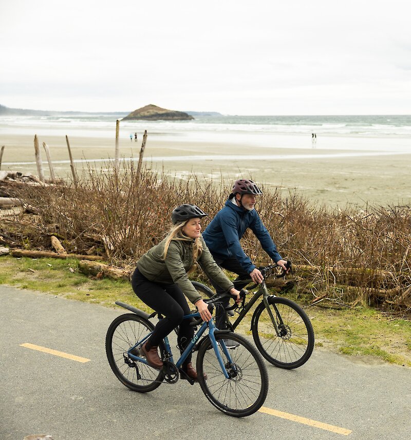 Two people road biking along the ʔapsčiik t̓ašii parallel to Incinerator Rock at Long Beach