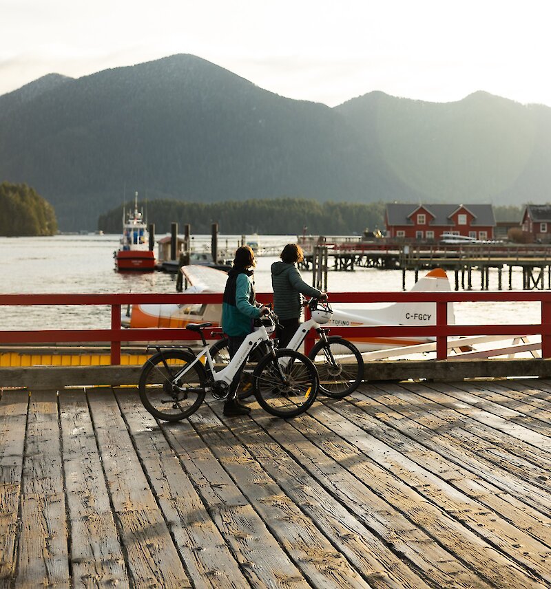 Two people standing beside electric bikes on First Street dock, overlooking the working harbour of Tofino.