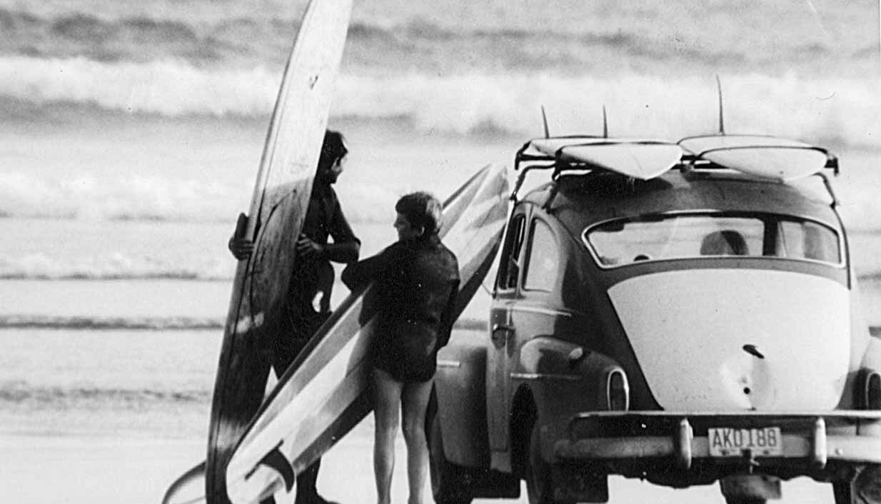 A black-and-white photo of a VW bug parked on the beach, two boards on the roof, two people, presumably father and son, holding two more boards as they look at the waves in the background.