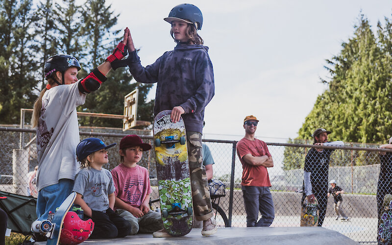 Two skaters in helmets high-fiving on the edge of the skate park, boards nearby and younger onlookers at Westcoast Triple Plank.