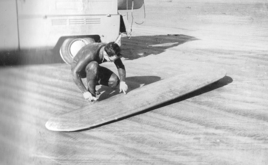 A black-and-white photo of Jim Sadler with his homemade surf board, prepping to go for a surf.