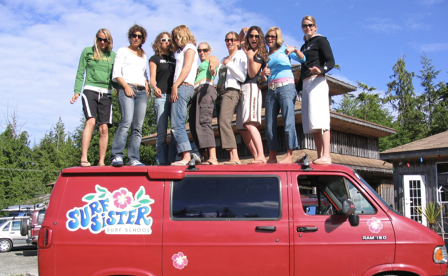A group of nine women waving shakas around in the early 2000's while standing on top of a red van with the Surf Sister logo.