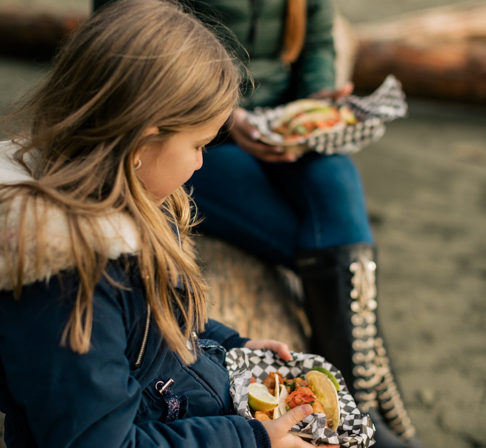 Child and adult eating tacos on a log at the beach