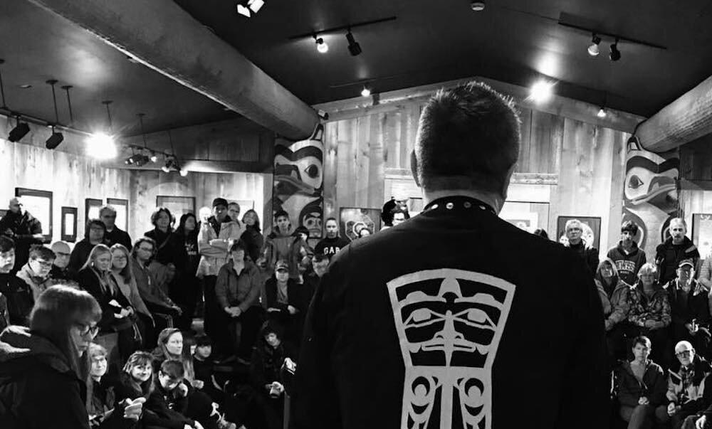 Man in Indigenous design vest, sitting next to drum and facing audience