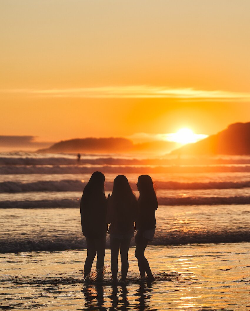 Three young women soak their feet in the water at sunset along Cox Bay.