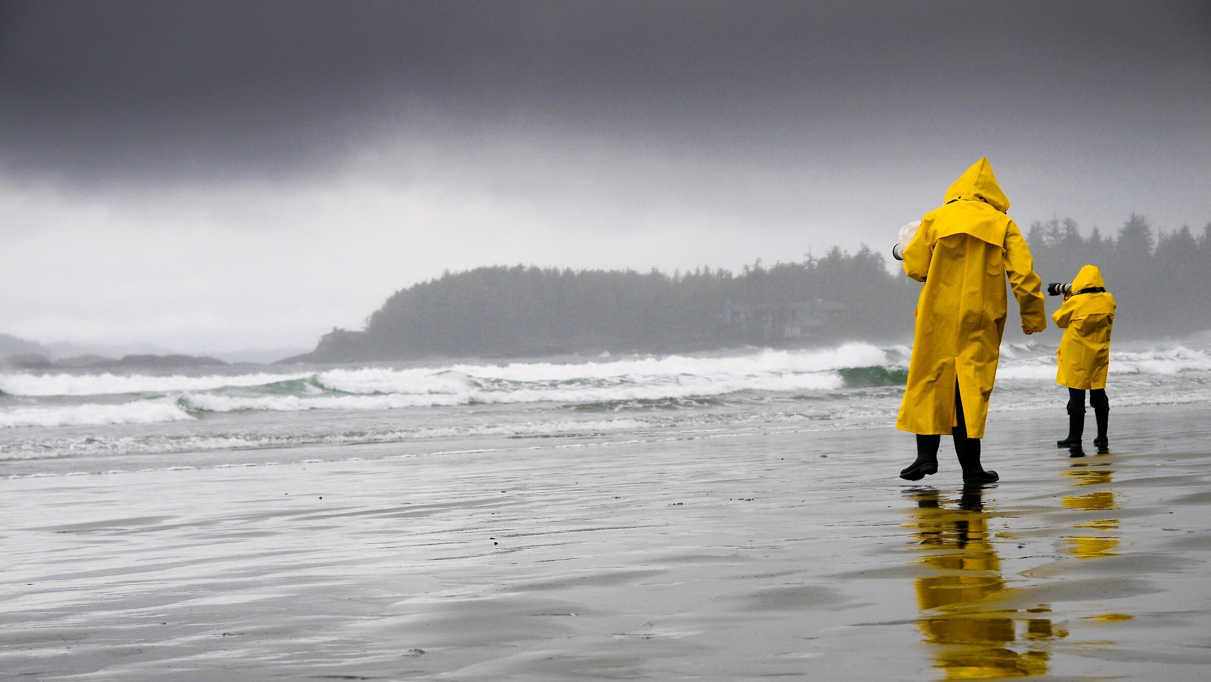 Two people on the beach in the rain, wearing yellow raincoats and taking photos of the storm