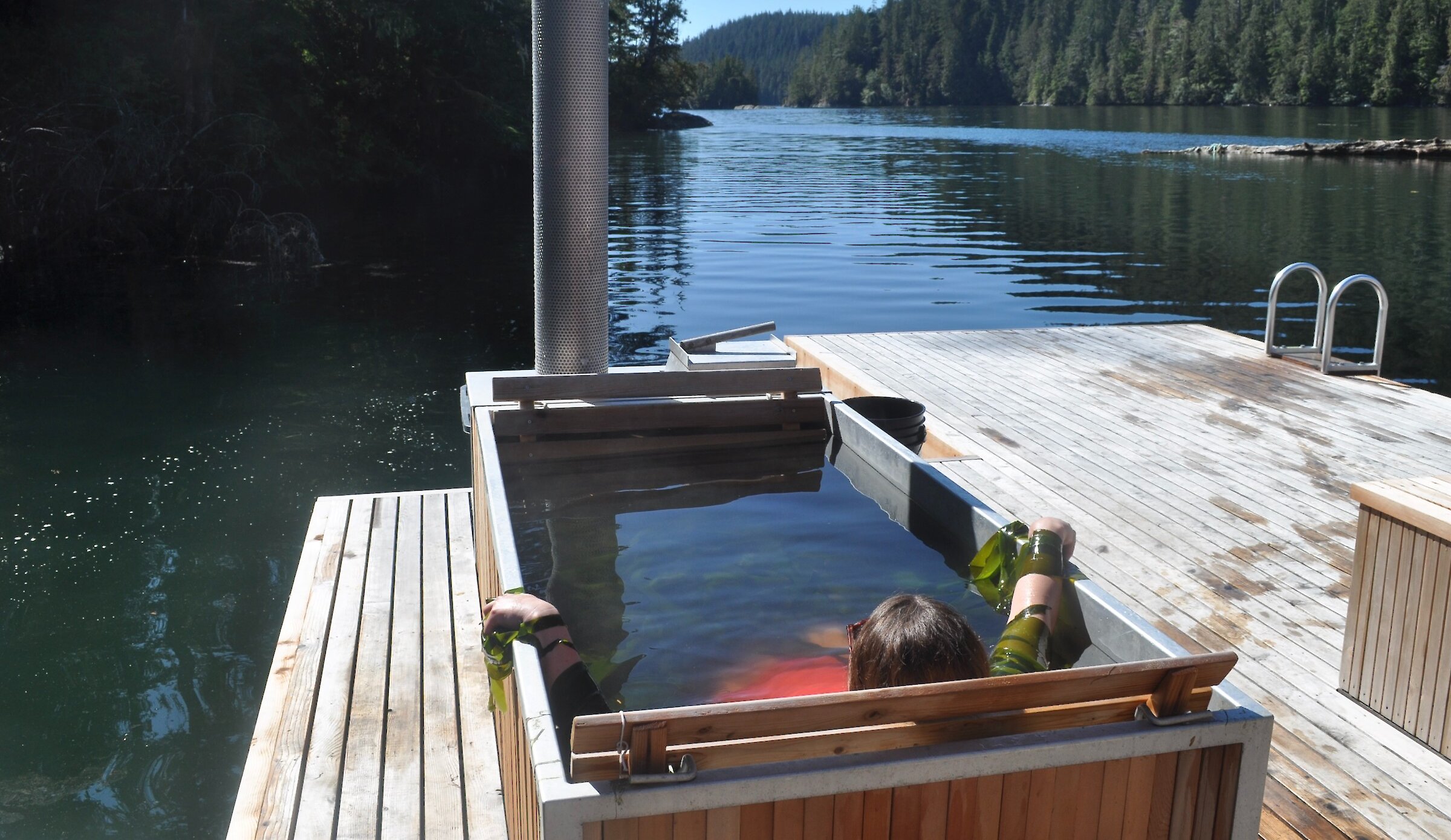 A woman soaks in a cedar-sided wood-fired tub on a floating dock with blue skies overhead