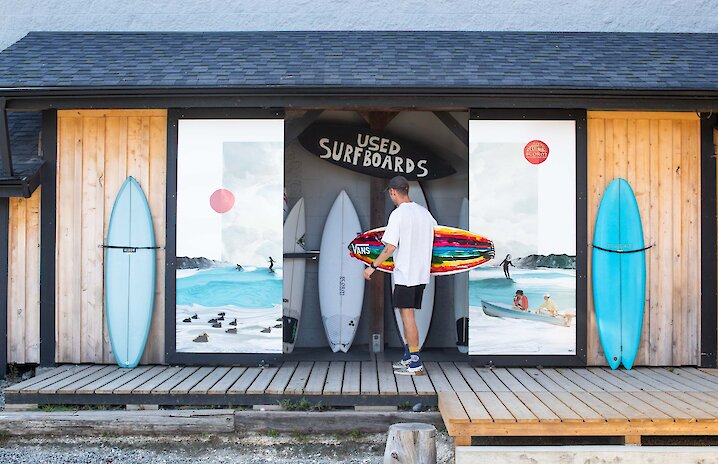 Person holding a surfboard outside of a surfshop with colourful murals on the doors