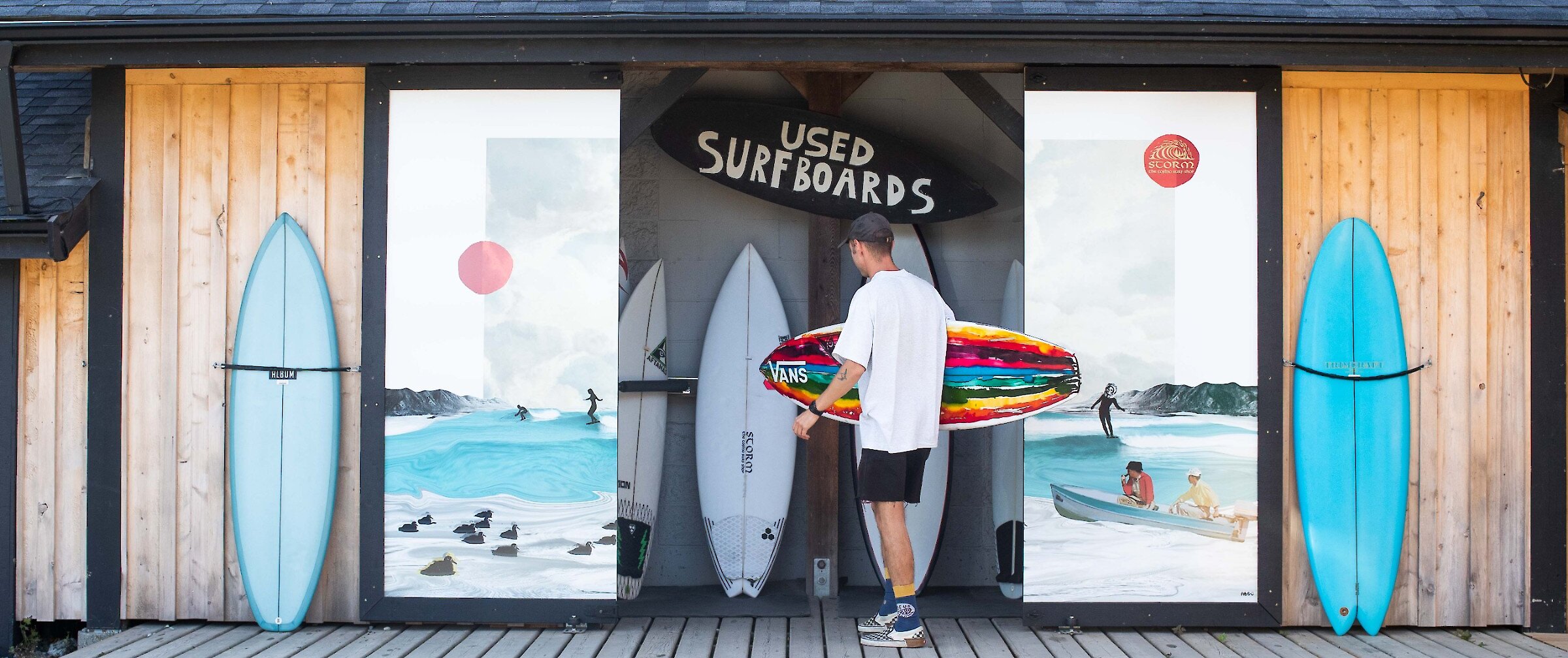 Person holding a surfboard outside of a surfshop with colourful murals on the doors