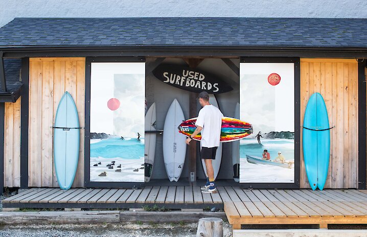 Person holding a surfboard outside of a surfshop with colourful murals on the doors