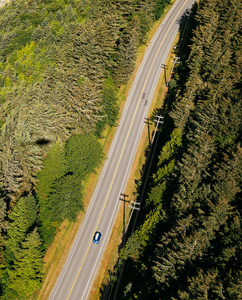 An aerial photo of a blue car with a surfboard on the roof driving a highway alongside forest and sandy beach through Pacific Rim National Park Reserve.