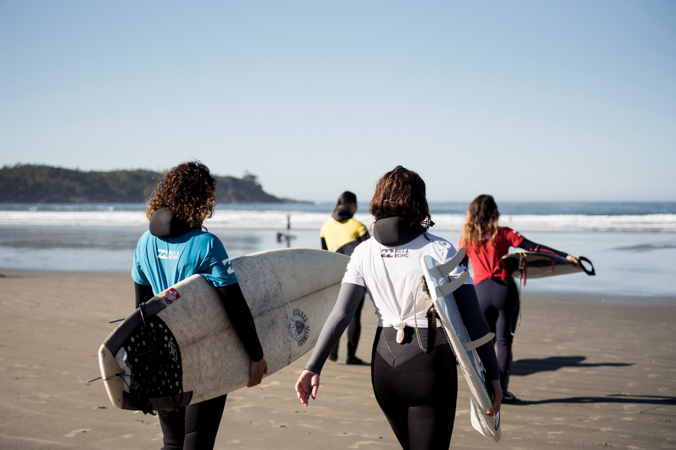 Four women surf competitors in differently coloured competition jerseys walking with their boards towards the ocean on Cox Bay.