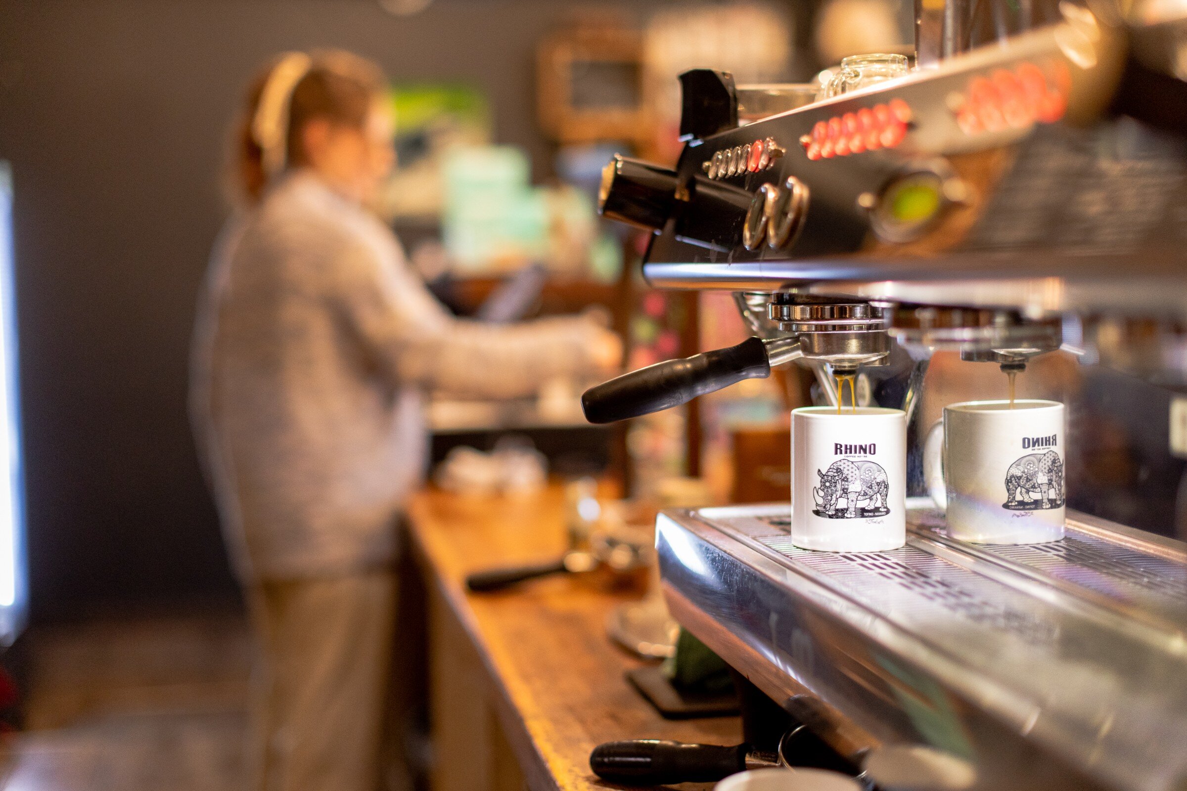 Close-up of a Rhino Coffeehouse mug sitting on a running espresso machine with a barista blurred in the background.