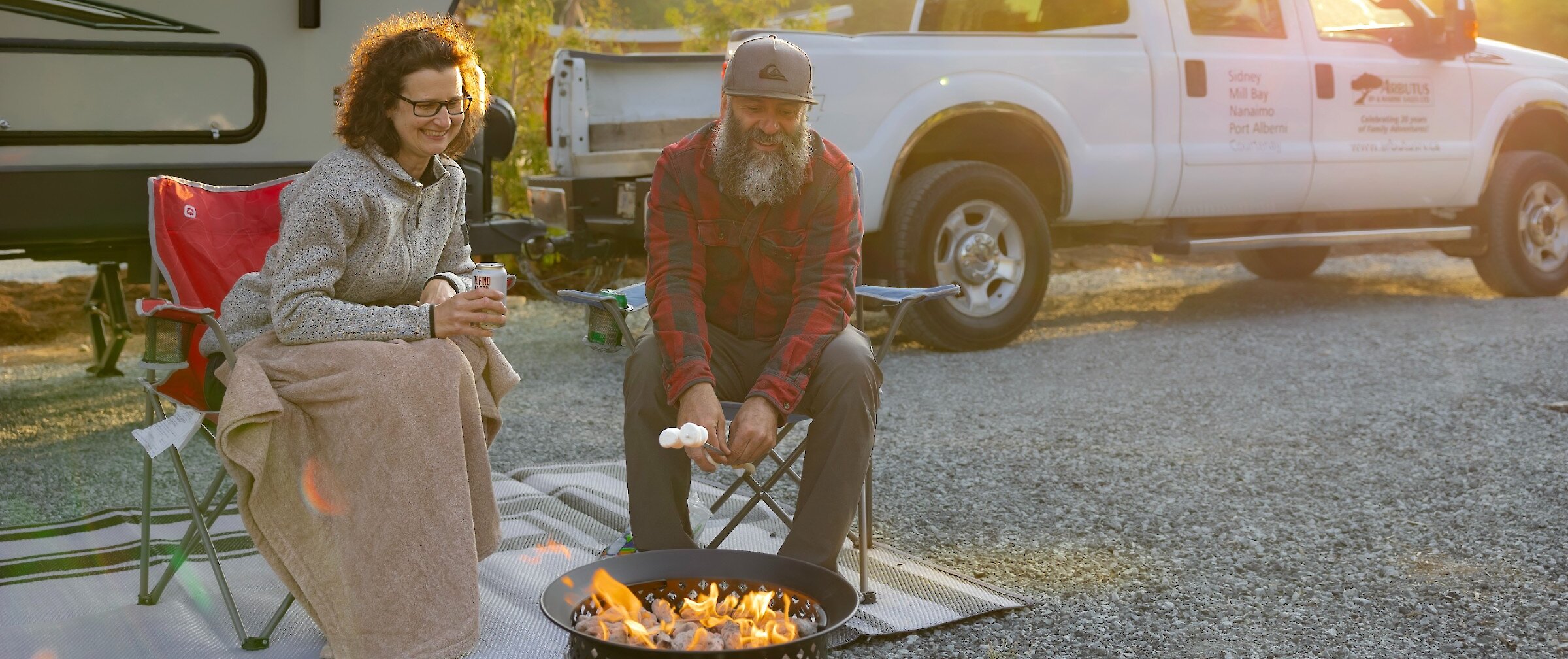 People in camping chairs roasting marshmallows over a fire with their camper in the background