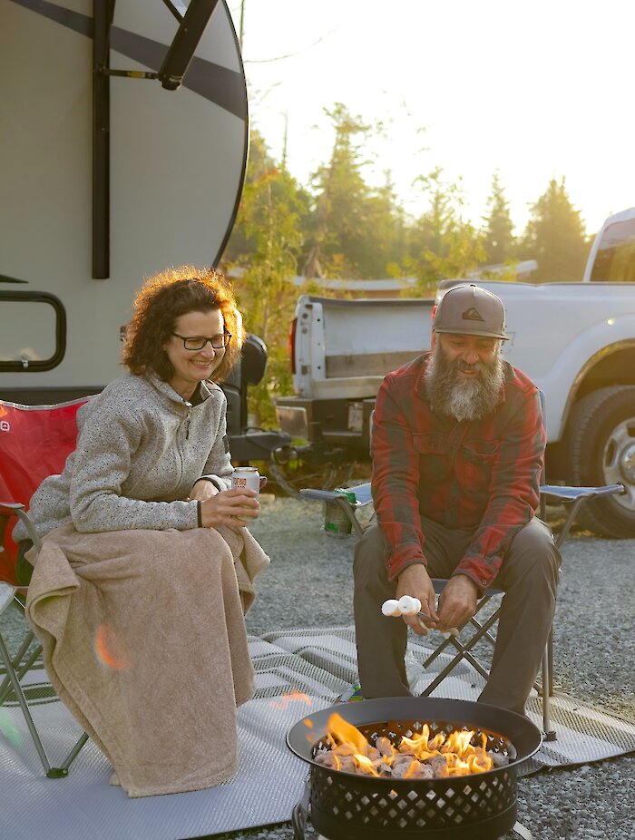 People in camping chairs roasting marshmallows over a fire with their camper in the background