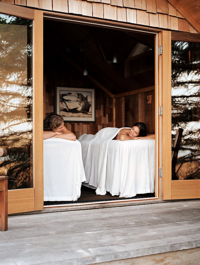 Two massage tables facing an open door out onto a patio overlooking the trees and ocean.