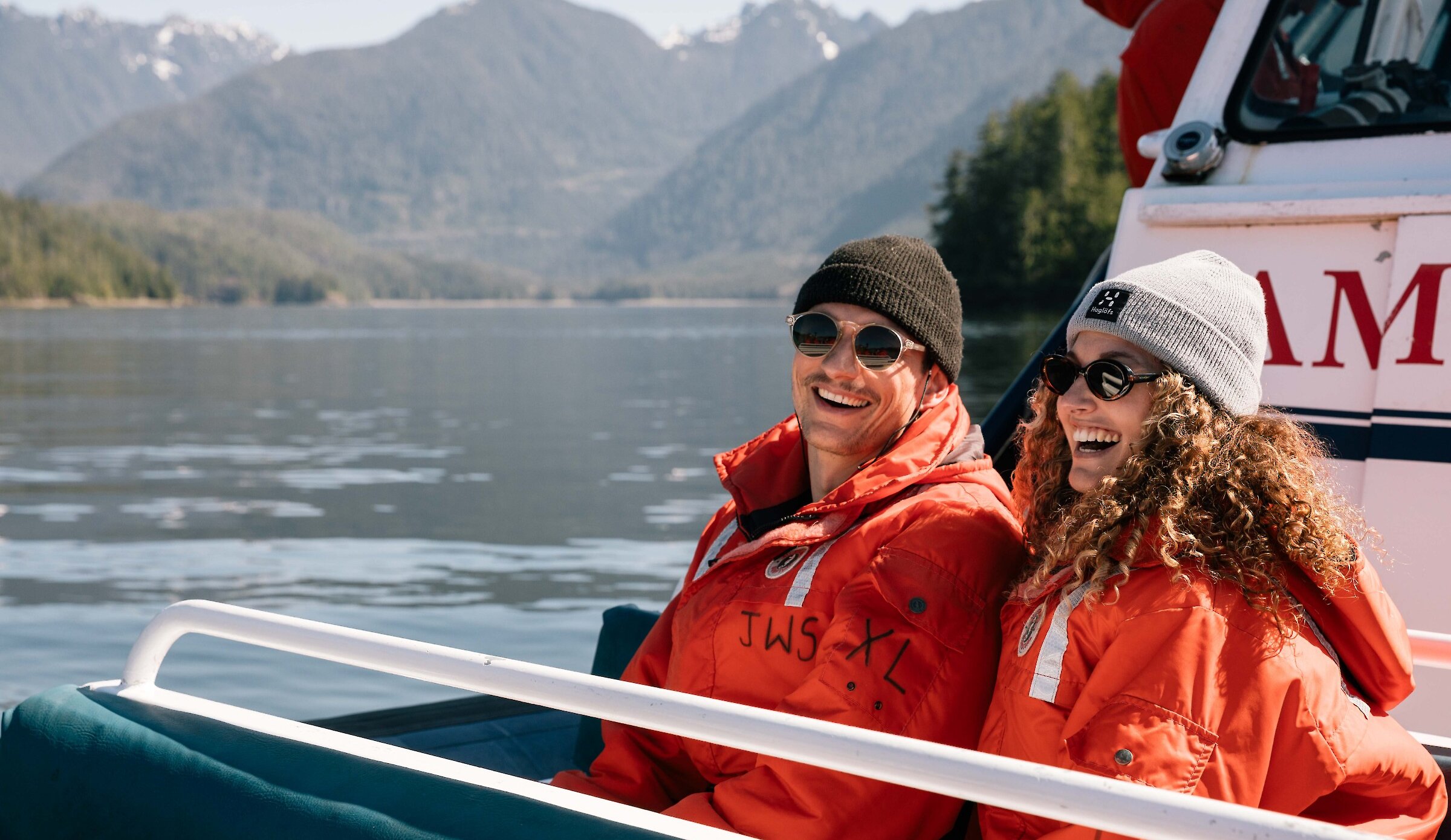 smiling man and woman in red floater suits on a Zodiac boat with hills in the background.