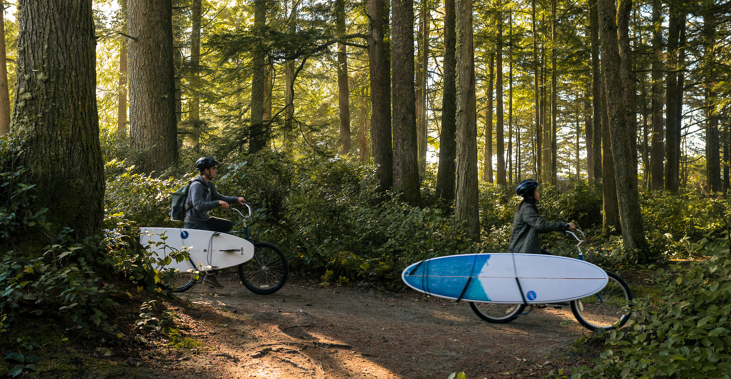 A couple riding through the forest with their bikes from Tofino Bike Company, on their way to their surf lesson with Pacific Surf Company.