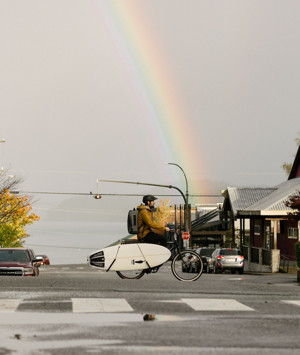 Person biking across a crosswalk with a surfboard and rainbow in the background