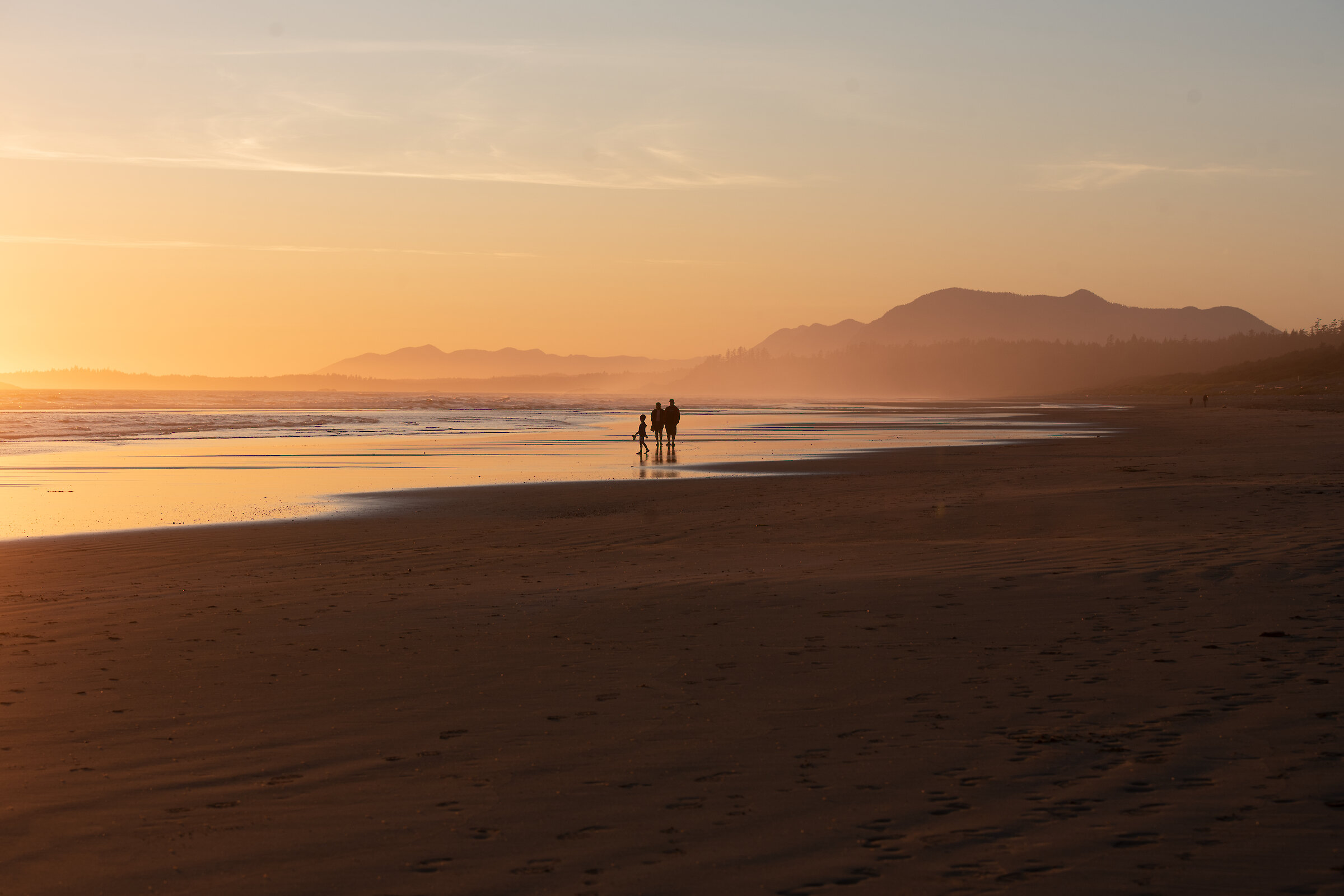 Two people and a child walking along Wickaninnish Beach at sunset