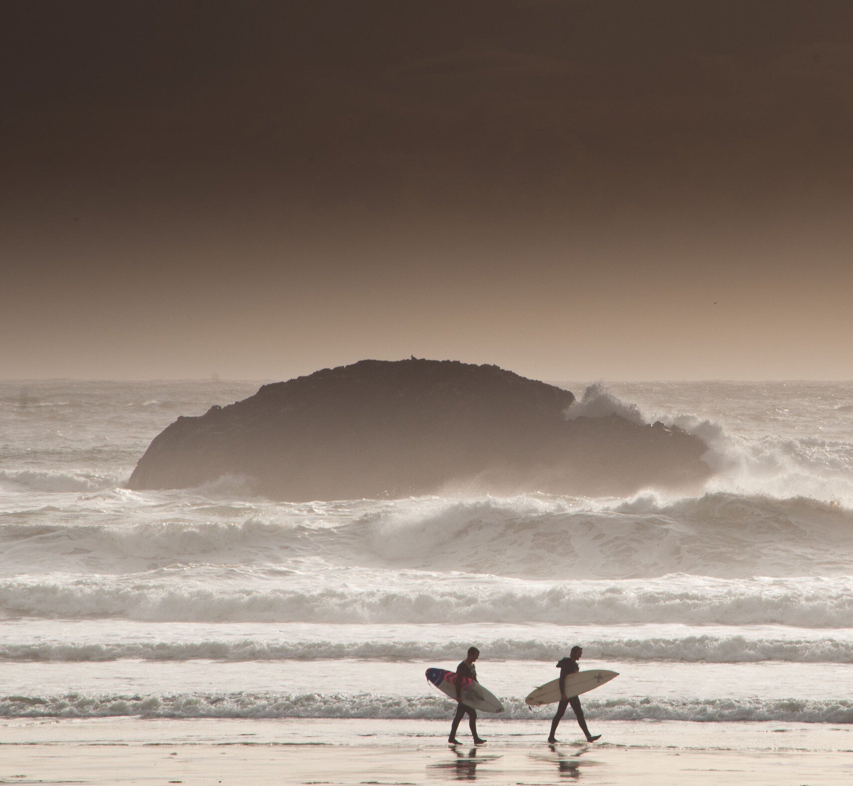 Two surfers walking along the beach with boards and surf and an island in the background