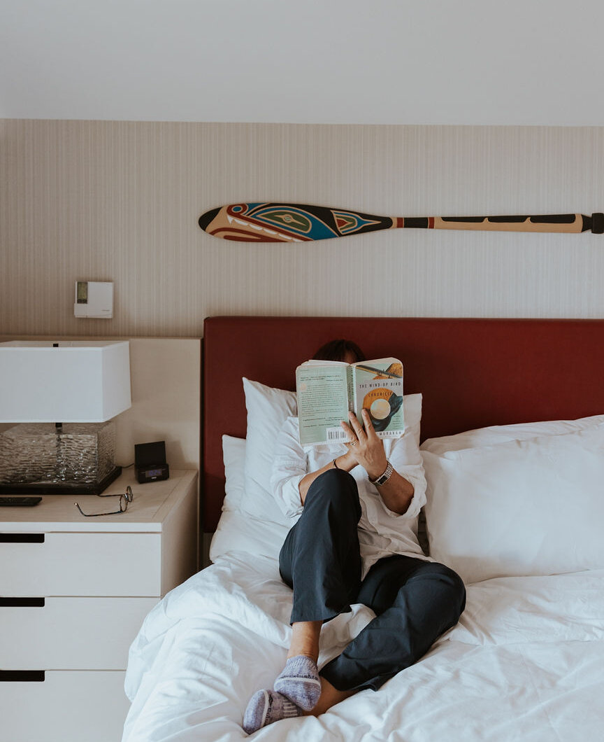 Woman relaxing on a bed in hotel room with a paddle as decor on the wall and reading a book