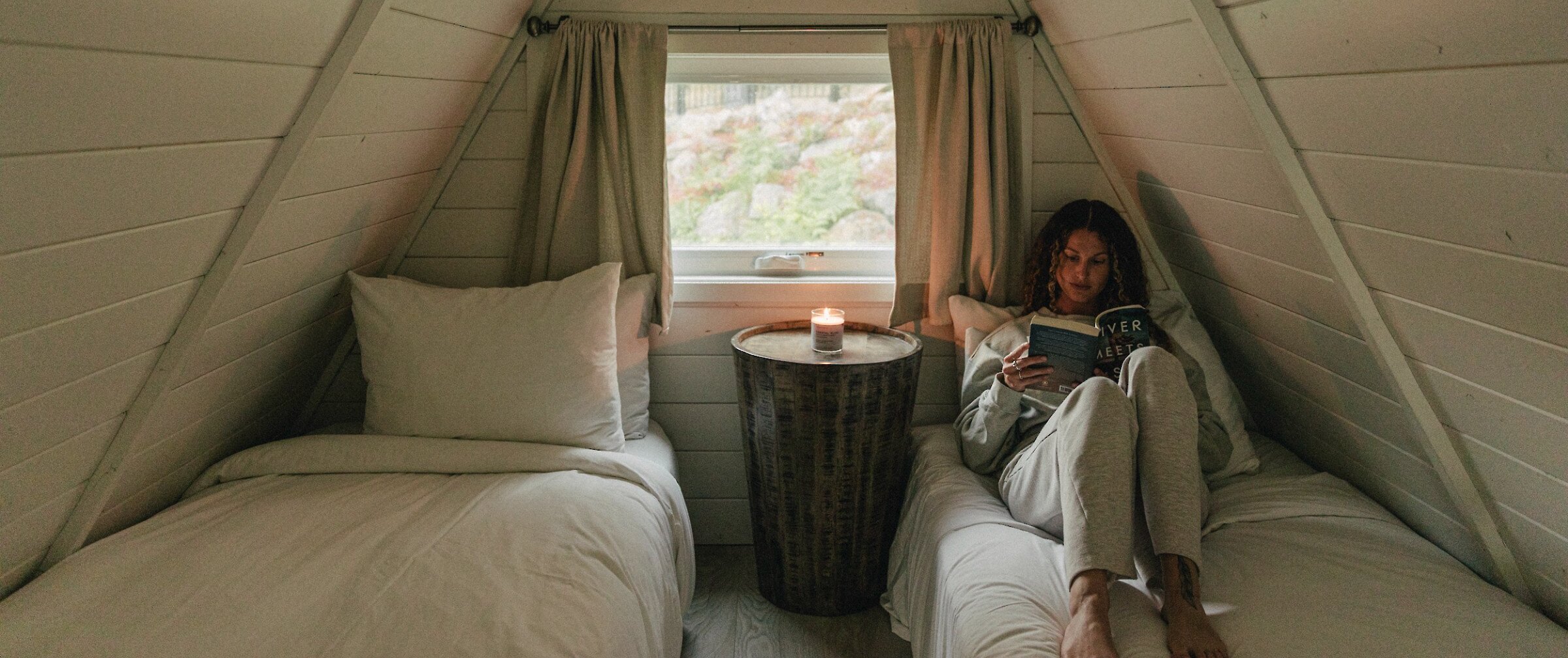 Person sitting on a bed reading a book next to a window with a lit candle in an A-frame cabin