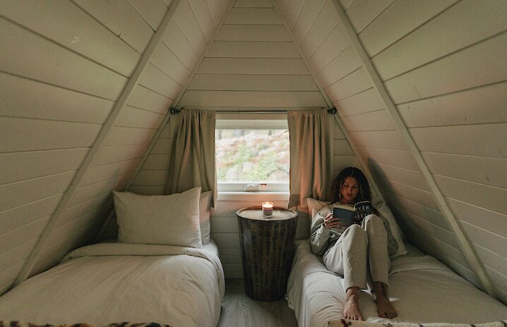 Person sitting on a bed reading a book next to a window with a lit candle in an A-frame cabin