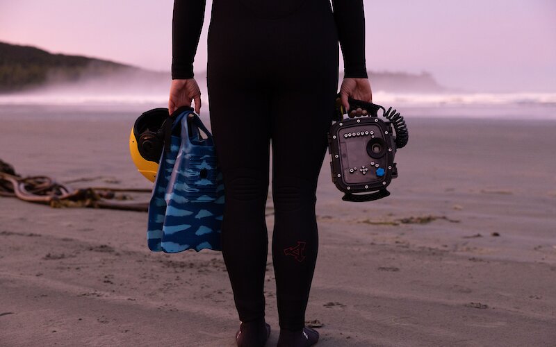 Person on the beach in a wetsuit holding fins, a camera and their helmet.