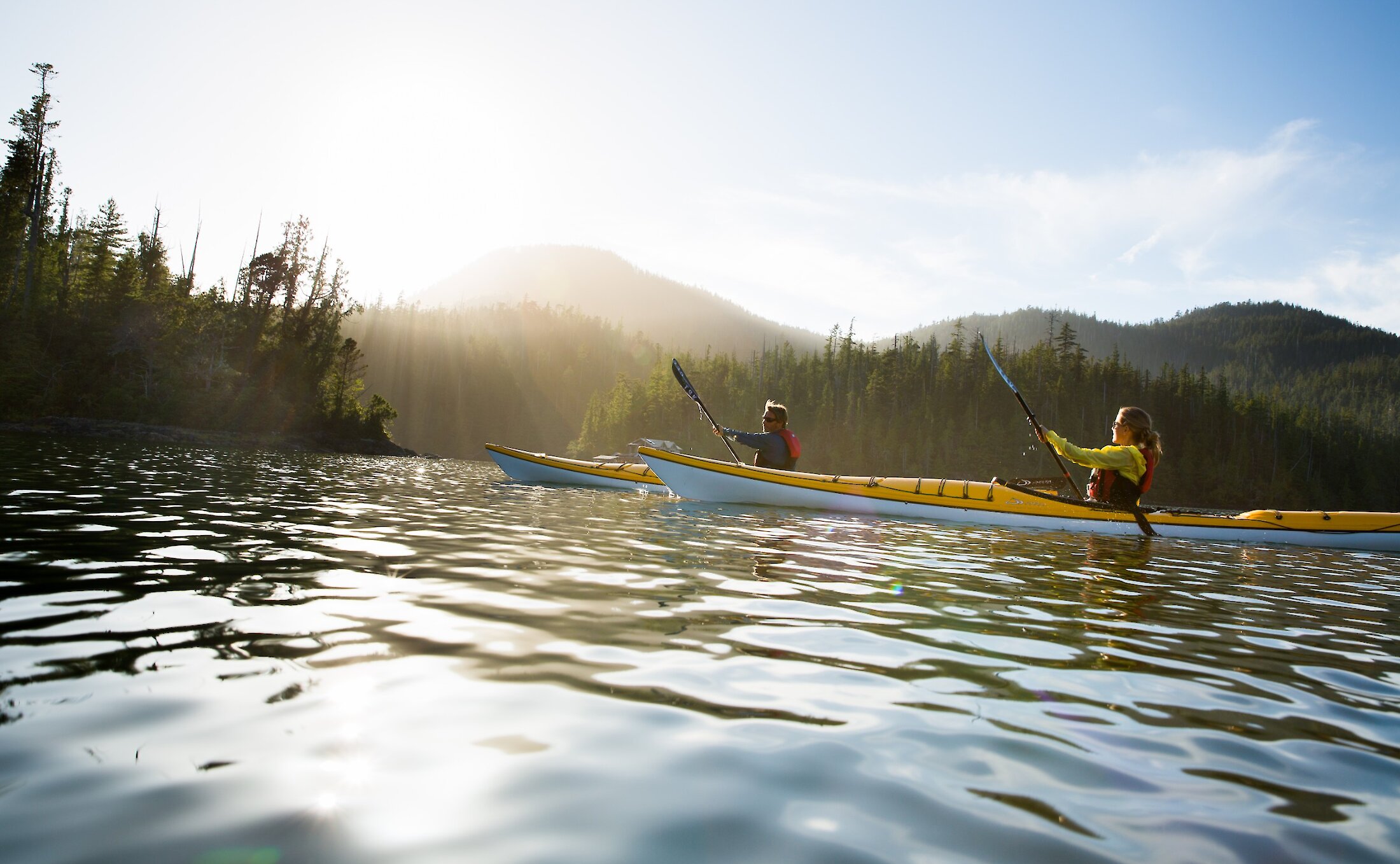 Two people kayaking with the misty mountains in the background on a sunny day.