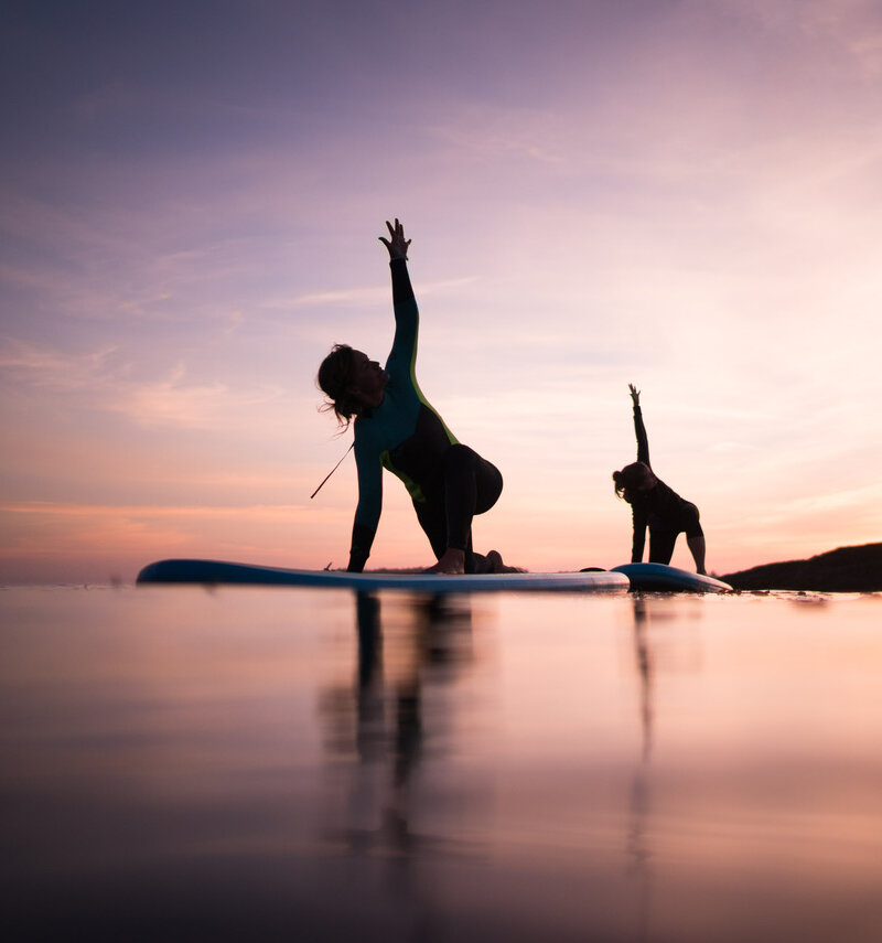 silhouettes of two people doing SUP yoga under a purple sunset