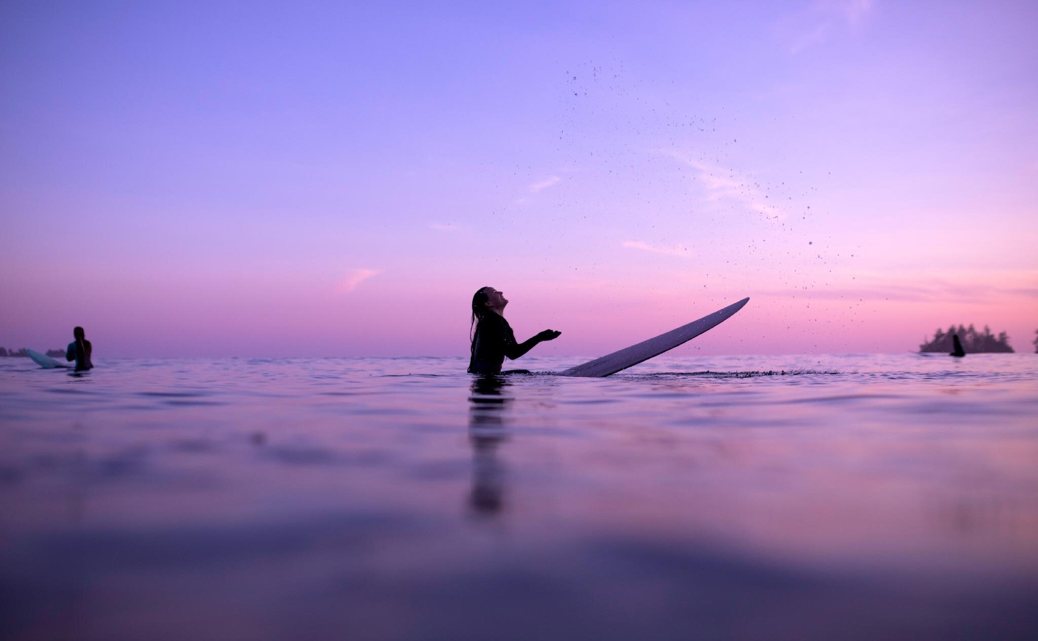 Purple skies at sunset with a surfer flipping water in her hands while sitting on her board in the ocean
