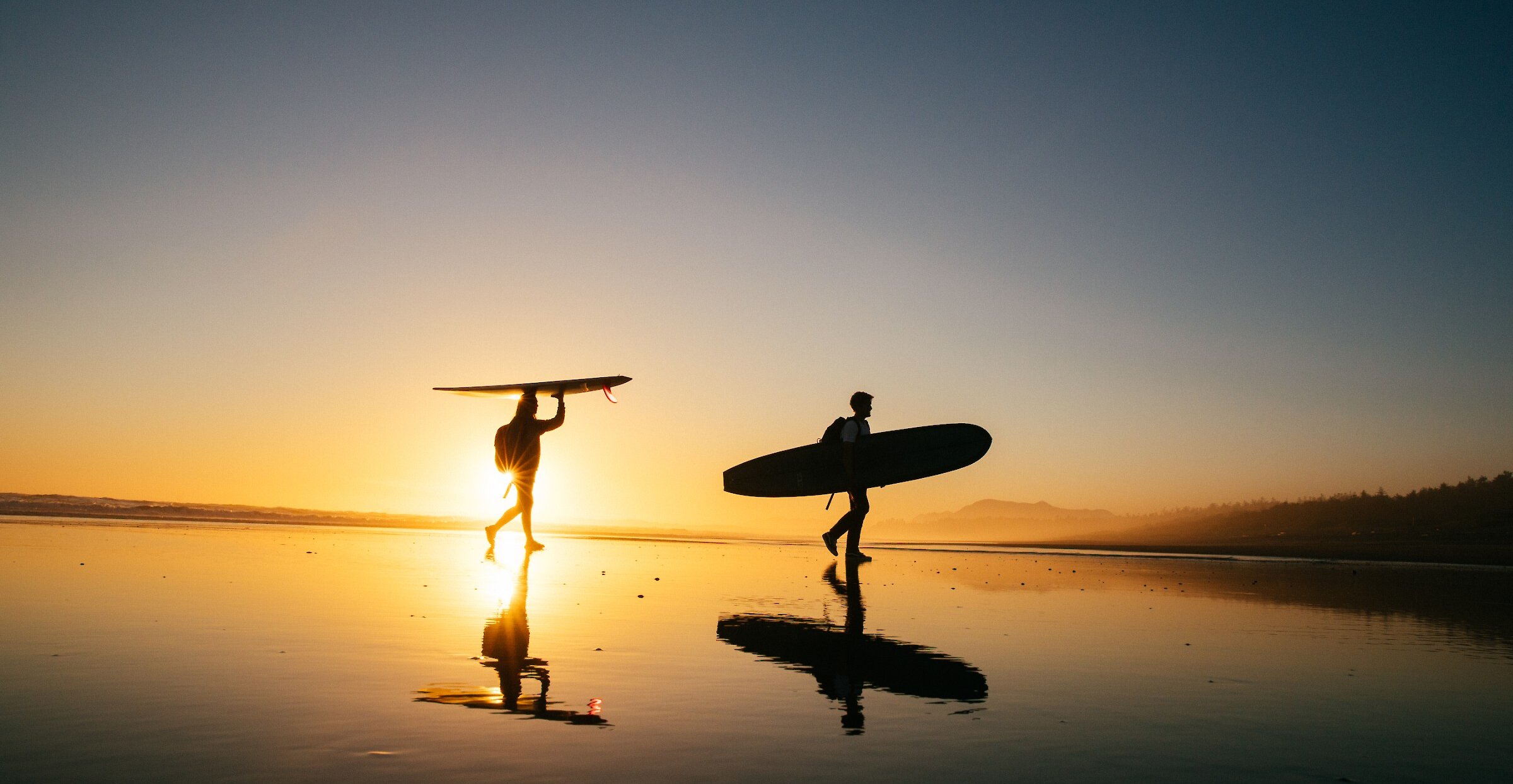 People walking on the glassy beach with their boards and the sunset in the background